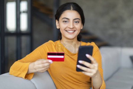 Foto de Mujer india alegre con tarjeta de crédito en la mano haciendo el pago en línea, hembra oriental feliz usando su teléfono móvil mientras está sentado en el sofá en el acogedor interior de la sala de estar en casa, espacio libre - Imagen libre de derechos