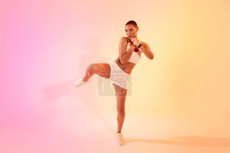 Téléchargez les photos : Une femme concentrée avec une tête rasée donne un puissant coup de pied dans une pose d'arts martiaux, portant un équipement sportif blanc sur fond rose et jaune mélangé. Fit, sport et soins de santé - en image libre de droit