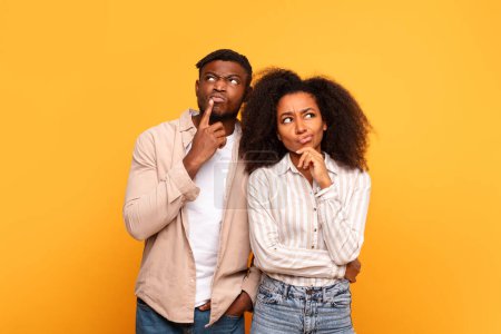Photo for Contemplative black man and woman in casual attire, with fingers on their cheeks, looking upwards in curiosity against plain yellow background - Royalty Free Image