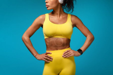 Photo for Unrecognizable athletic african american lady with fitness tracker watch holding hands on hips, standing against blue studio background, wearing yellow fitwear. Cropped shot - Royalty Free Image