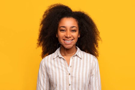 Fröhliche junge schwarze Dame mit lockigem Haar und breitem Lächeln, in lässig gestreiftem Hemd, vor leuchtend gelbem Hintergrund stehend
