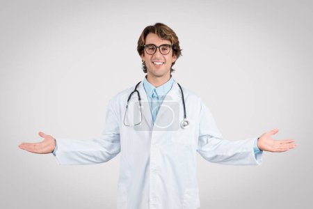 Photo for Smiling male doctor with glasses and stethoscope opens his arms wide in welcoming gesture, standing against a light gray background - Royalty Free Image