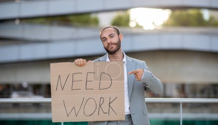 Photo for Young man in suit holding Need Work sign, illustrating the search for employment opportunities in tough job market, upset male standing outdoors against modern office building, copy space - Royalty Free Image