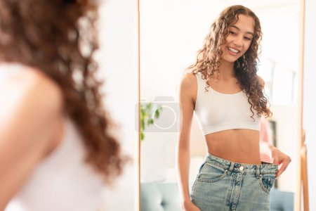 Photo for Caucasian teen girl smiling as she compares her waist size at home, wearing big jeans to show weight loss achievement, looking at her mirror reflection at home. Healthy slimming concept - Royalty Free Image