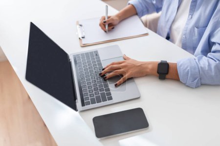 Photo for Close-up of a professional multitasking with a laptop and taking notes, featuring a smartwatch and a smartphone on a clean, white desk in a modern workspace, cropped, top view - Royalty Free Image