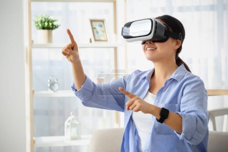 Foto de Técnicos modernos y entretenimiento. Emocional joven mujer asiática en gafas VR sentado en el sofá en casa, jugando videojuegos interactivos, explorando la realidad aumentada, tocando el espacio de copia - Imagen libre de derechos