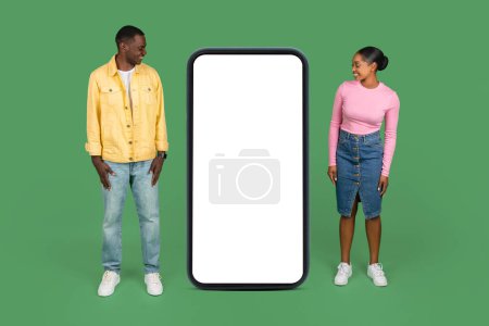 Photo for Curious young black man and woman looking at huge phone with white blank screen. Smiling millennial african american couple check nice online offer or mobile app, mockup, copy space, green background - Royalty Free Image