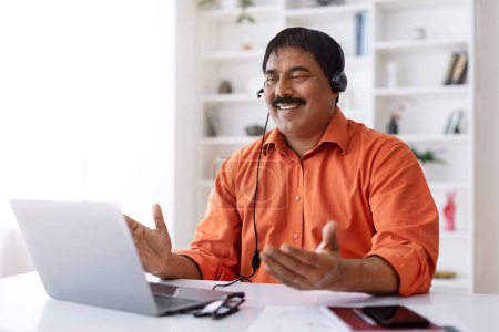 Téléchargez les photos : Homme d'affaires indien mature ont un appel vidéo avec le client ou le partenaire d'affaires, assis au bureau, en utilisant un ordinateur portable et un casque d'écoute, travaillant à partir du bureau à la maison, réunion en ligne avec des collègues, espace de copie - en image libre de droit