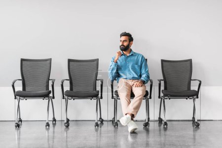Photo for Thoughtful professional indian man seated alone among empty chairs in stark office setting, lost in contemplation or awaiting an important event - Royalty Free Image