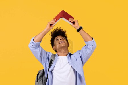 Téléchargez les photos : Jeune étudiant noir ludique créant un toit au-dessus de sa tête avec un livre rouge, regardant vers le haut avec un sourire rêveur sur fond de studio jaune - en image libre de droit
