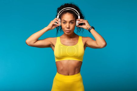 Photo for Fit black lady in yellow sportswear using wireless headphones while training, standing over blue backdrop in studio, symbolizing healthy active lifestyle. Workout motivation concept - Royalty Free Image
