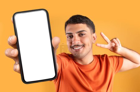 Téléchargez les photos : Jeune homme brésilien insouciant montrant geste de paix et smartphone avec écran blanc, ayant une humeur ludique, gars positif posant sur fond de studio orange, recommandant application mobile - en image libre de droit