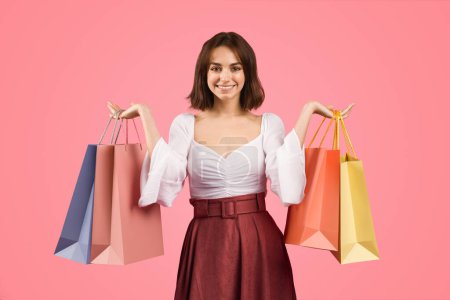 Photo for Smiling young european woman in a stylish white blouse and maroon skirt, exuding happiness and confidence, holding colorful shopping bags on a vibrant pink background, studio - Royalty Free Image