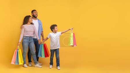 Photo for Family Shopping. Black Preteen Boy Pointing Finger Aside Showing Something To His Parents While They Standing Together With Shopper Bags Over Yellow Studio Background, Panorama With Copy Space - Royalty Free Image