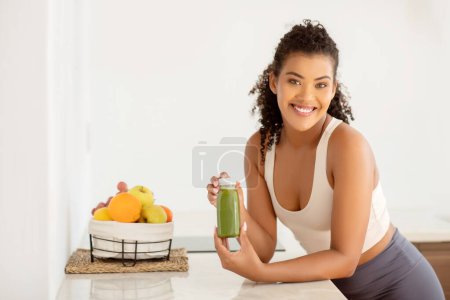 Photo for Fit woman in activewear holds nutritious green smoothie made of fresh fruits and vegetables, enjoying morning ritual for wellness and weight loss at home kitchen interior - Royalty Free Image