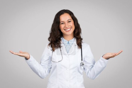 Photo for Joyful female doctor with stethoscope outstretching both arms, palms up in balancing gesture, wearing white coat against grey backdrop - Royalty Free Image
