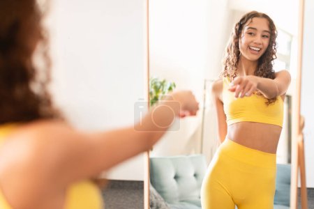 Foto de Sonriente adolescente adolescente en ropa deportiva apunta a la reflexión en el espejo en casa, el uso de ropa de vestir amarillo listo para el entrenamiento de fitness, disfrutar de una vida deportiva saludable. Cuerpo positivo y autoaceptación - Imagen libre de derechos