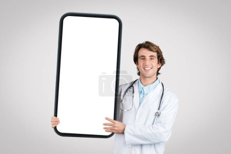 Médico masculino sonriente con bata de laboratorio sosteniendo un smartphone gigante con pantalla en blanco para maqueta, de pie sobre fondo gris, lugar para anuncio médico