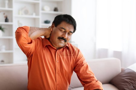 Photo for Suffering middle aged indian man feeling neck pain, sitting on couch in cozy living room at home. Chronic injury, muscle inflammation strain and backache concept, copy space - Royalty Free Image