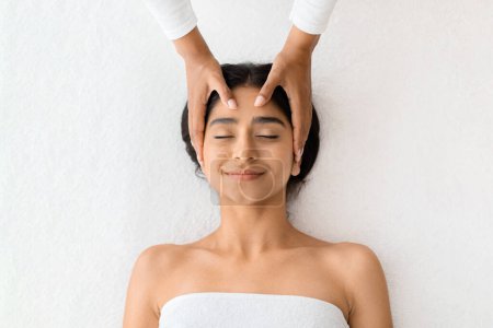 Photo for A content young indian woman smiles in bliss as skilled hands gently massage her forehead, symbolizing relaxation and self-care in a soothing environment, top view, white background - Royalty Free Image