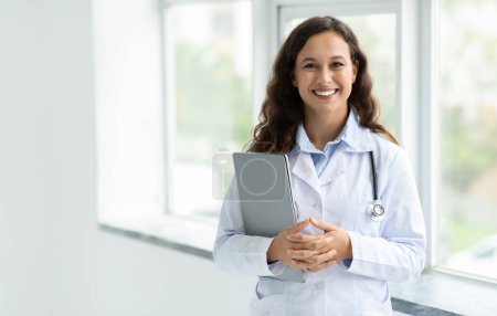 Cheerful young caucasian woman in white medical coat doctor standing by window at clinic hospital, holding laptop computer, online consultation with patient, copy space. Telehealth, remote healthcare