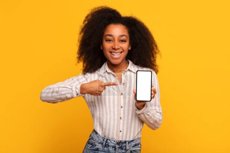 Entzückte junge schwarze Dame zeigt auf Smartphone mit leerem Bildschirm, perfekt für die Anzeige einer App oder Website-Werbung, vor leuchtend gelbem Hintergrund