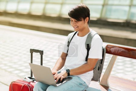 Photo for Asian guy freelancer independent contractor sitting on bench, working on laptop while waiting for train at station. Remote employee chinese man checking email on computer at airport, copy space - Royalty Free Image