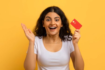 Photo for Excited emotional pretty young indian woman wearing white t-shirt showing her red plastic bank credit card, gesturing and grimacing, yellow background. Easy banking, fast loan - Royalty Free Image