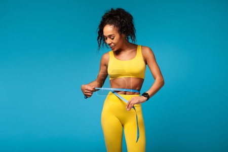 Foto de Mujer afroamericana fitness con cinta métrica, comprobando el tamaño de la cintura después de adelgazar, posando en ropa deportiva amarilla sobre fondo azul del estudio, simbolizando la pérdida de peso y el cuidado del cuerpo - Imagen libre de derechos