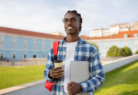 Téléchargez les photos : Heureux étudiant afro-américain se tient debout tenant ordinateur portable et tasse de café, mélangeant loisirs et apprentissage à l'extérieur au parc universitaire. Apprentissage en ligne et approche numérique moderne de l'éducation - en image libre de droit