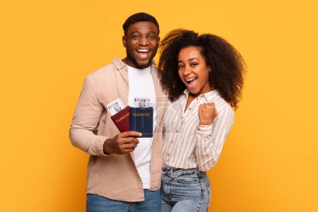 Excited young black couple holding passports with boarding passes, showing winning gestures, happy about their upcoming trip on yellow background