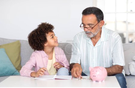 Téléchargez les photos : Économies familiales. Grand-père joyeux Calculer de l'argent avec son petit-fils, prendre des notes assis avec une tirelire, apprendre les finances à la maison. Concept d'abondance et d'investissements financiers - en image libre de droit