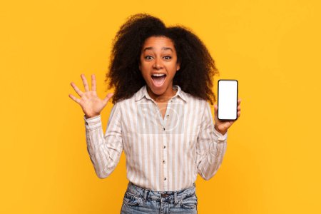 Photo for Joyful young black lady with curly hair excitedly presents smartphone with blank screen, gesturing with her other hand, against vibrant yellow backdrop - Royalty Free Image