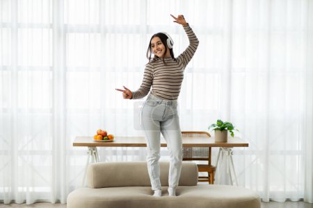 Joyful young woman with headphones dancing on a beige couch, throwing her arms up in a playful manner, in a bright airy room with a sheer curtain background. Fun with music at home