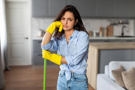 Foto de Mujer cansada en camisa azul y guantes amarillos apoya su mejilla en su mano, apoyándose en la fregona en la cocina moderna, tomando una pausa de las tareas domésticas - Imagen libre de derechos