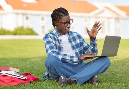 Téléchargez les photos : Heureux étudiant noir étudiant sur ordinateur portable saluant bonjour, assis sur le campus universitaire parc herbe en plein air, immergé dans l'apprentissage en ligne lors d'une conférence vidéo ou virtuelle à distance - en image libre de droit