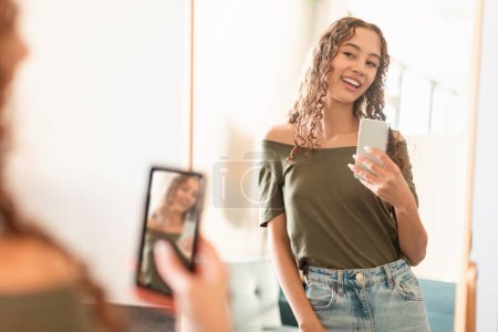 Téléchargez les photos : Jeune adolescente faisant du selfie via smartphone posant joyeusement près du miroir à la maison, capturant moment de style de vie des jeunes pour les médias sociaux à l'intérieur. Blogging et gadgets concept. Concentration sélective - en image libre de droit