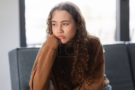 Photo for Upset young caucasian teen girl sitting alone on her sofa indoors, leaning chin on hand, reflecting boredom and signs of teenage apathy, portrait shot. Modern youth mental health concept - Royalty Free Image