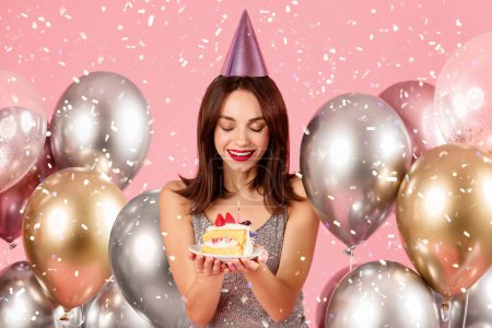 Téléchargez les photos : Délicieuse femme caucasienne millénaire dans un chapeau de fête et robe scintillante admirant une tranche de gâteau d'anniversaire, avec des ballons d'hélium et une douche de confettis en arrière-plan - en image libre de droit