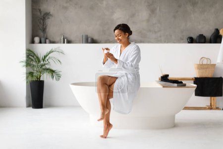 Photo for Skincare. Happy Young African American Woman Applying Cream In Bathroom, Smiling Black Female Moisturizing Skin After Bath, Sitting On Luxury Bathtub, Having Beauty Day At Home, Copy Space - Royalty Free Image