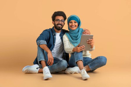 Téléchargez les photos : Divertissement en ligne. Heureux jeune couple musulman utilisant la tablette numérique ensemble tout en étant assis sur le sol sur fond beige dans Studio, joyeux conjoints arabes naviguant sur Internet sur le gadget moderne - en image libre de droit