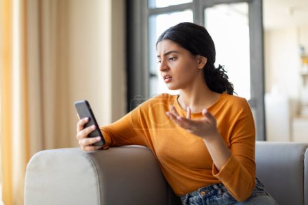 Téléchargez les photos : Frustrée jeune femme indienne regardant son smartphone avec un geste d'incrédulité, confuse femelle orientale avec téléphone portable à la main assise sur le canapé dans le salon à la maison, lisant un message désagréable - en image libre de droit
