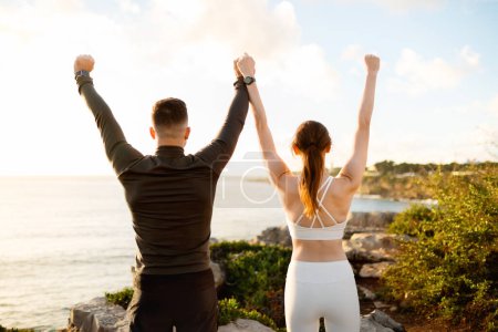 Foto de Vista posterior del hombre y la mujer levantando sus brazos en la victoria, celebrando el logro de la aptitud en el acantilado costero con un impresionante horizonte marítimo - Imagen libre de derechos