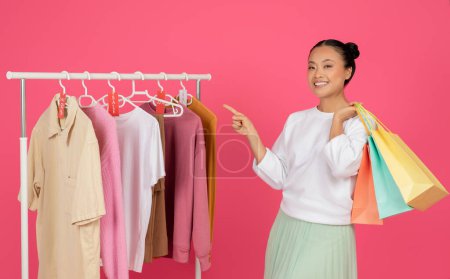 Photo for Happy Asian Woman Holding Shopping Bags And Pointing At Rack With Trendy Clothes, Cheerful Korean Female Advertising Seasonal Sales, Enjoying Making Purchases, Standing On Pink Studio Background - Royalty Free Image