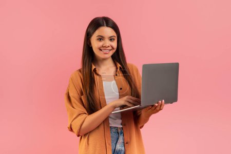 Téléchargez les photos : Adolescente confiante tenant un ordinateur portable et souriant à la caméra, belle adolescente portant une tenue décontractée debout sur fond de studio rose, représentant l'éducation en ligne ou le travail à distance - en image libre de droit
