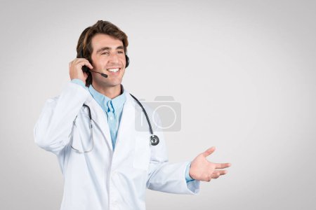 Foto de Médico masculino sonriente que usa auriculares y bata blanca de laboratorio que tiene una conversación alegre por teléfono, indicativo de comunicación de telesalud - Imagen libre de derechos