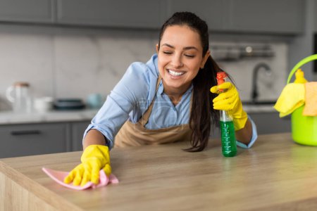 Photo for Cheerful woman in yellow gloves and an apron cleans kitchen counter with pink cloth and green spray bottle, smiling as she works, closeup - Royalty Free Image