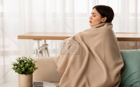 Foto de Mujer joven frustrada deprimida envuelta en manta sentada en el sofá en casa, vista lateral, espacio para copiar. Dama fría congelándose en el apartamento sin calor durante el invierno - Imagen libre de derechos