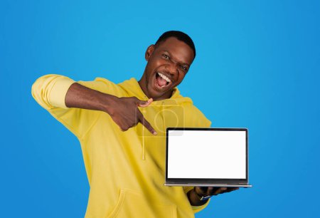 Téléchargez les photos : Un homme noir extatique dans un sweat à capuche jaune pointe avec excitation sur un écran d'ordinateur portable avec un écran vierge, parfait pour les maquettes ou les présentations, sur un fond bleu vif - en image libre de droit