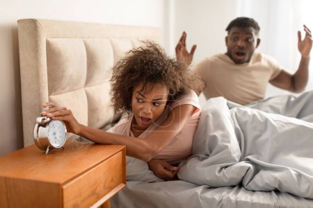 Photo for Oversleeping. Shocked black couple missed ringing alarm clock, stressed about being late to work, lying in bed in modern bedroom at home in the morning. Time management issue - Royalty Free Image